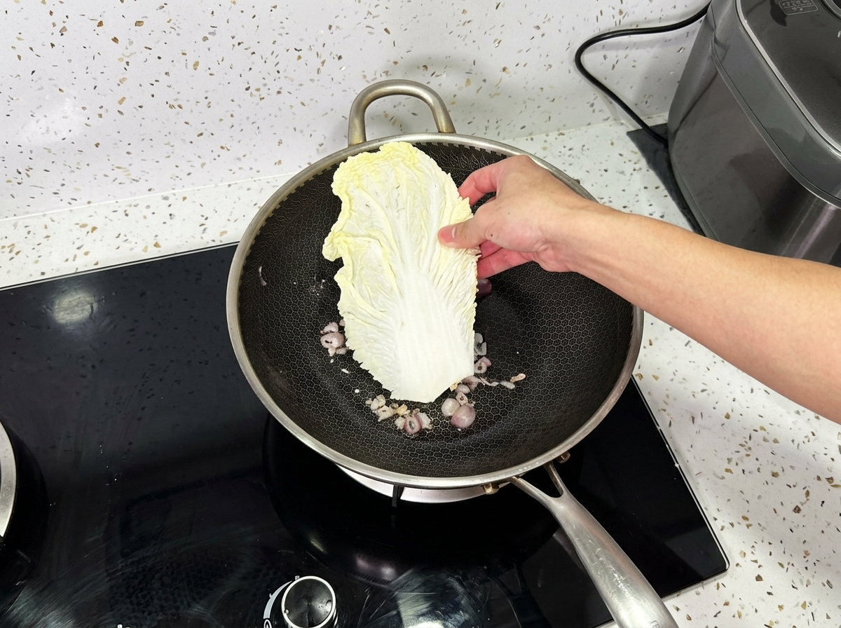 炒めた玉ねぎが入った中華鍋に、白菜の大きな葉を片手で入れようとしているところ。