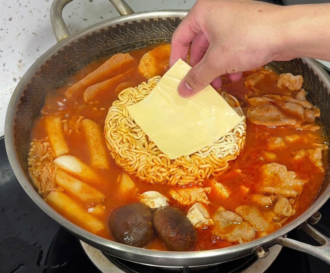 湯気の立つ部隊チゲの中で、ラーメンの麺の上に黄色いチーズのスライスがとろけているところ。