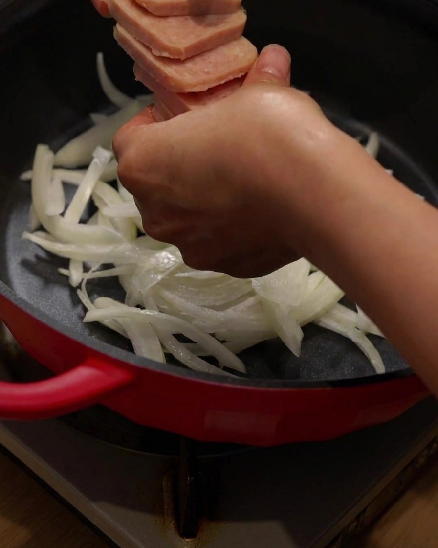 赤い調理鍋の中で、玉ねぎの上にスライスしたランチョンミートとベーコンを並べている様子。