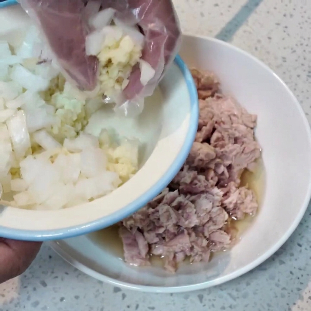 プラスチックの手袋をつけた手が、小さなボウルから刻んだ玉ねぎとニンニクを、ツナが入った大きなボウルに移しているところ。