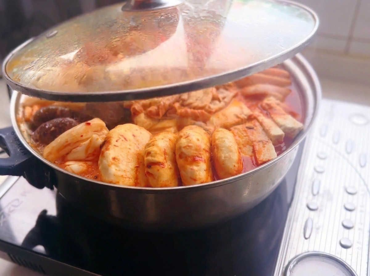 IHコンロの上の真っ赤な辛口スープに、スライス肉、豆腐、キノコ、トック、餃子などの具材がきれいに並べられた金属製の鍋から、ガラス蓋が持ち上げられている様子。