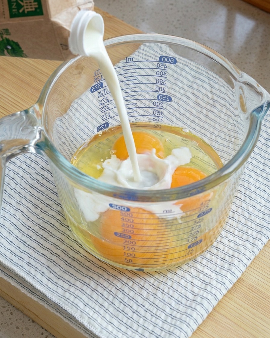 3個の生卵が入った大きなガラスの計量カップに牛乳を注ぐ様子。