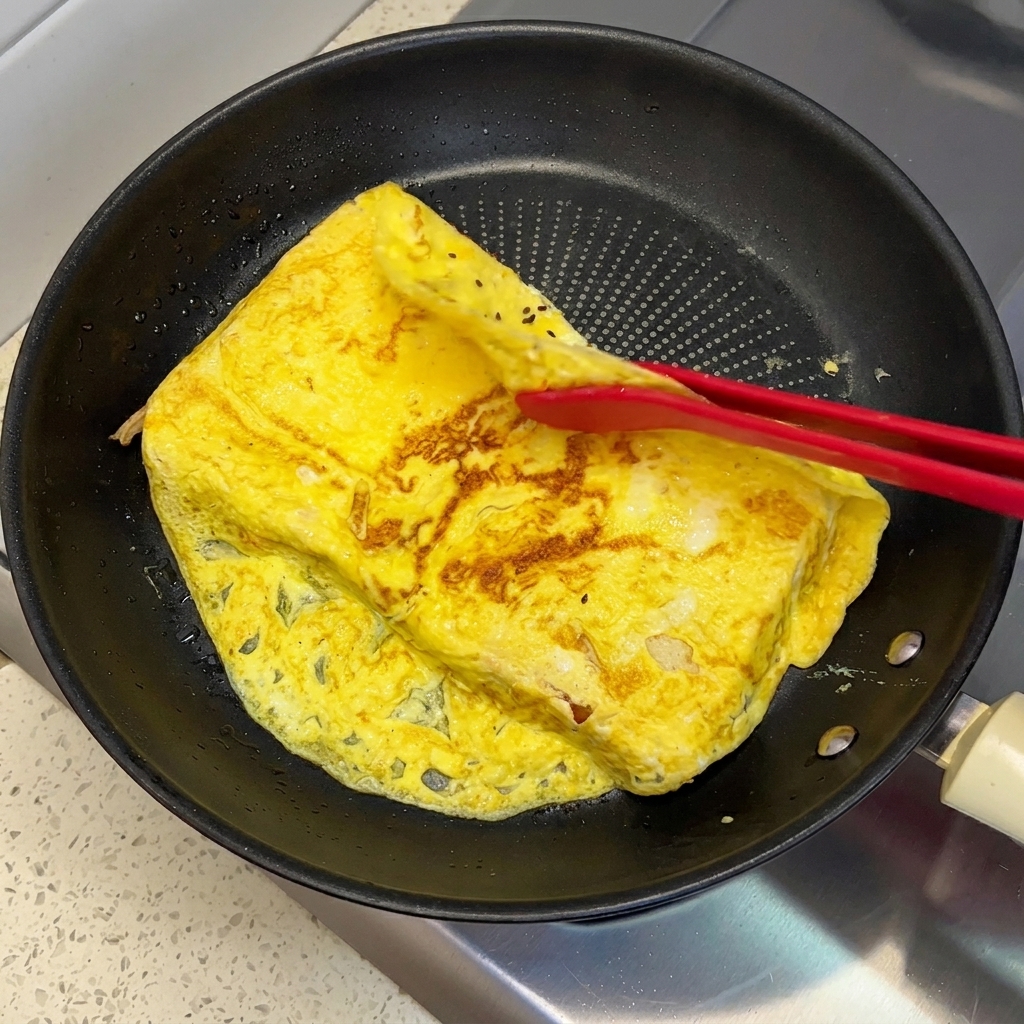 赤色のシリコン製トングで、調理された卵オムレツのはみ出た端をフライパン内の2枚の食パンの上に折りたたんでいるところ。