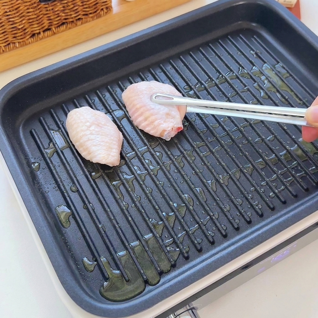 トングを使って、油を引いたリブ付きグリルパンに生の手羽先が並べられている様子。