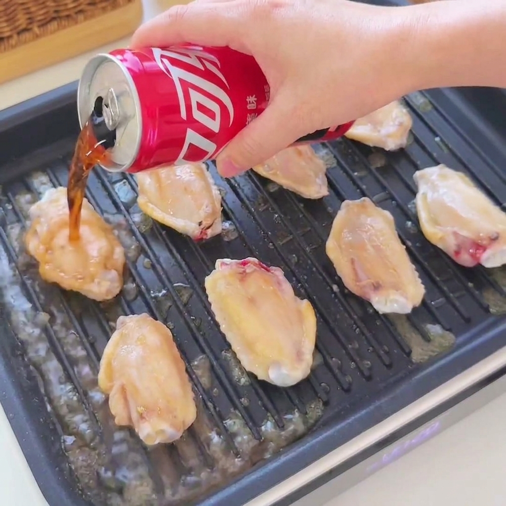 黒いグリルパンでジューシーに焼ける手羽先の上に、赤い缶からコーラと醤油が注がれている様子。
