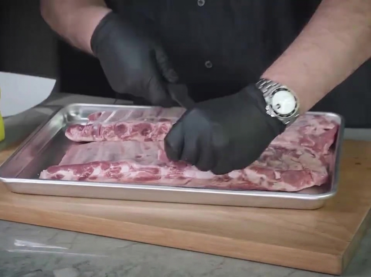 スチール製トレイの上で生肉のセントルイスリブの余分な脂肪をナイフで処理している様子。