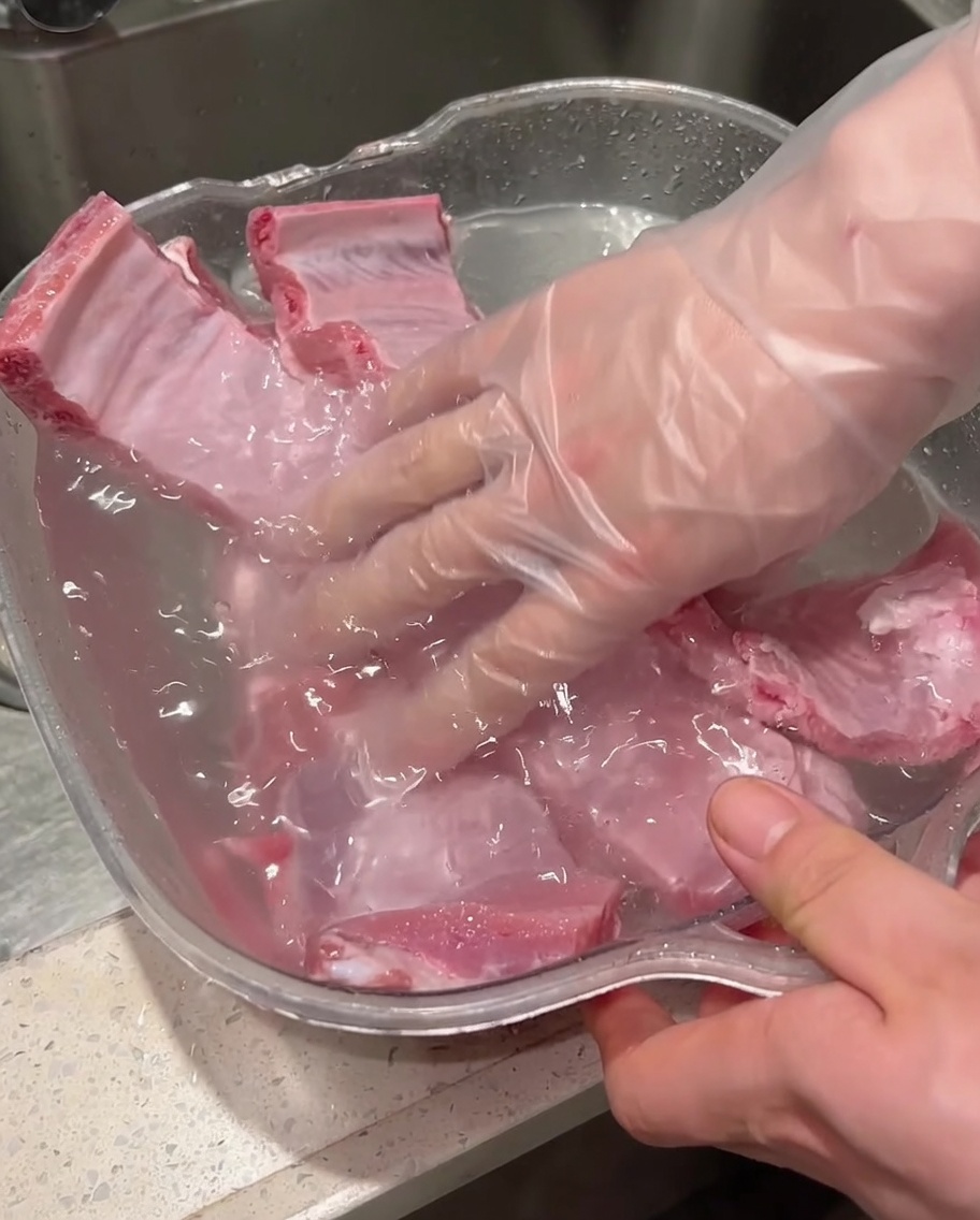 手袋を着用した手が水を入れた容器の中で生の豚スペアリブをきれいに洗い流している様子。