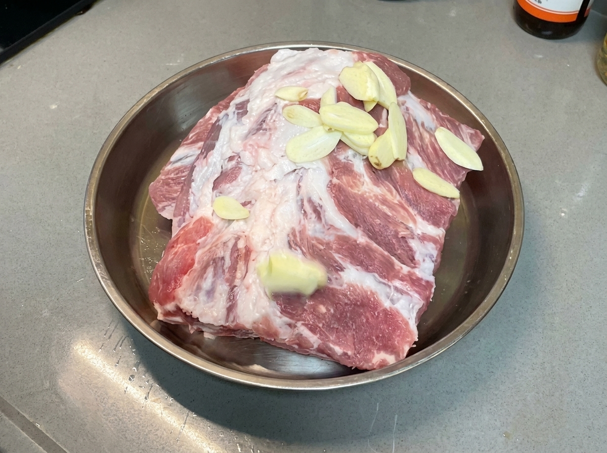 金属製のトレイに入った生ポークリブの上に、にんにくのスライスが均等に散らされている様子。
