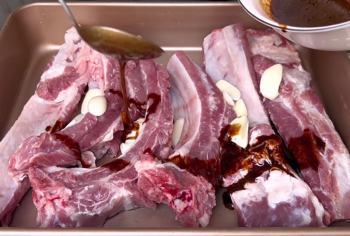白いボウルから、生肉の豚スペアリブとニンニクスライスが入ったバットへ、醤油ベースの濃いマリネ液を注いでいる様子。