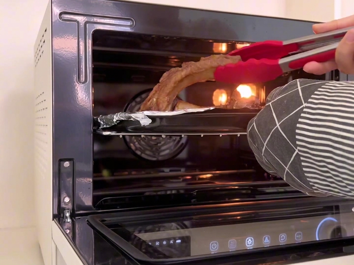赤いシリコン製のキッチントングが、オーブントレイの中で焼き上がった豚スペアリブの向きを裏返している様子。