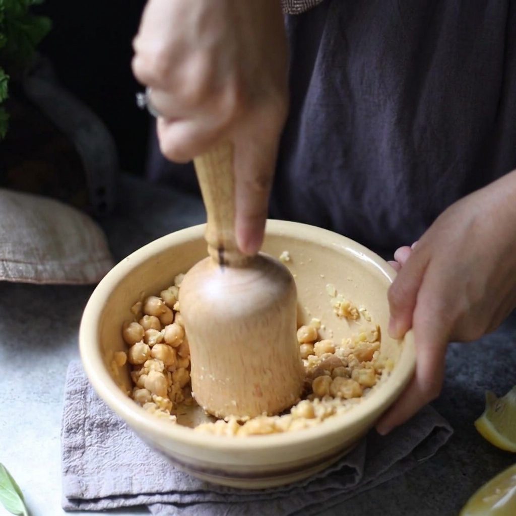 黄色い陶器のボウルの中で、木製のすりこぎ棒を使って茹でたひよこ豆を手作業で潰している様子。