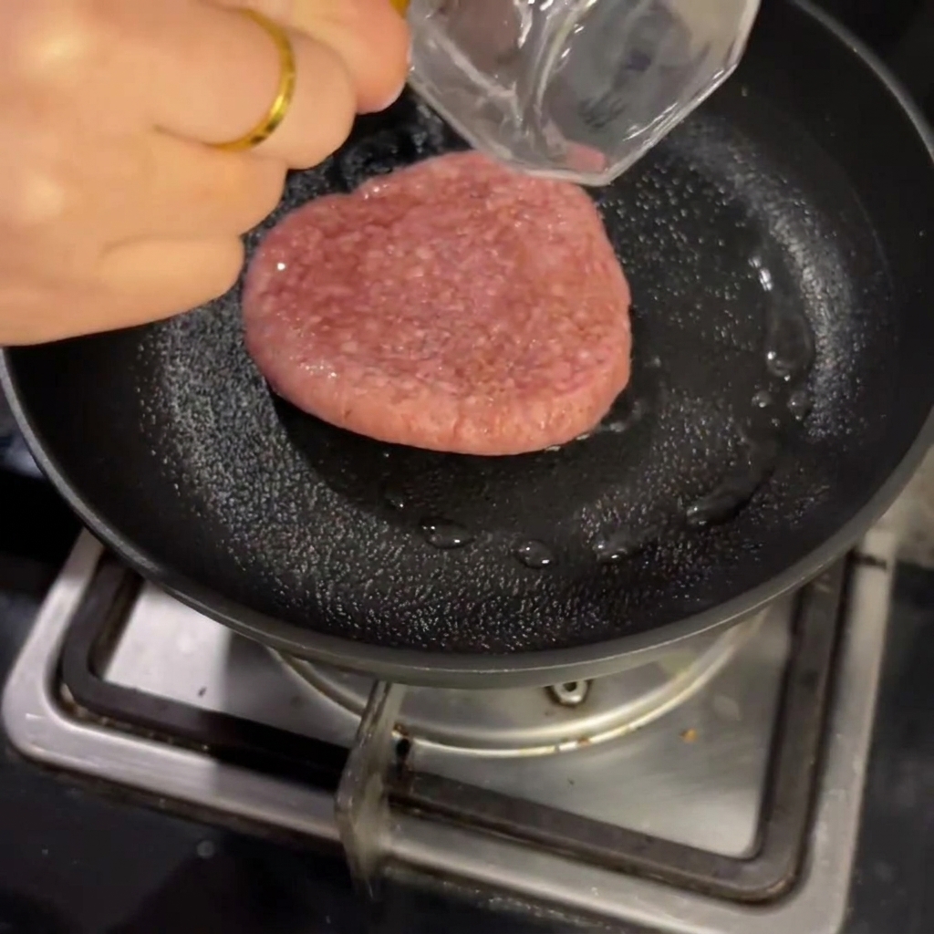 フライパンで生の牛パティを少量の水とともに蒸し焼きにする様子。