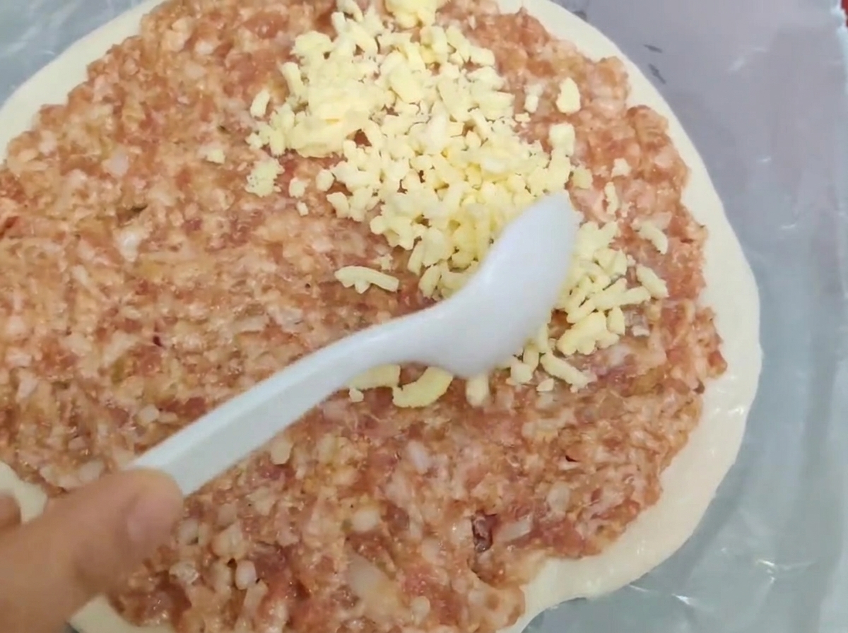 丸い生地の上にスプーンで生のひき肉とシュレッドチーズを広げている様子。