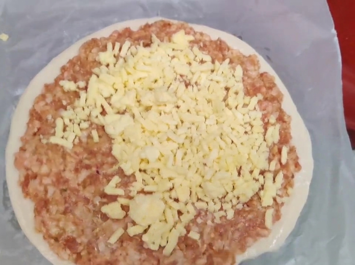 ネギ餅の上の生のひき肉に、黄色いシュレッドチーズがたっぷりと乗せられている様子。