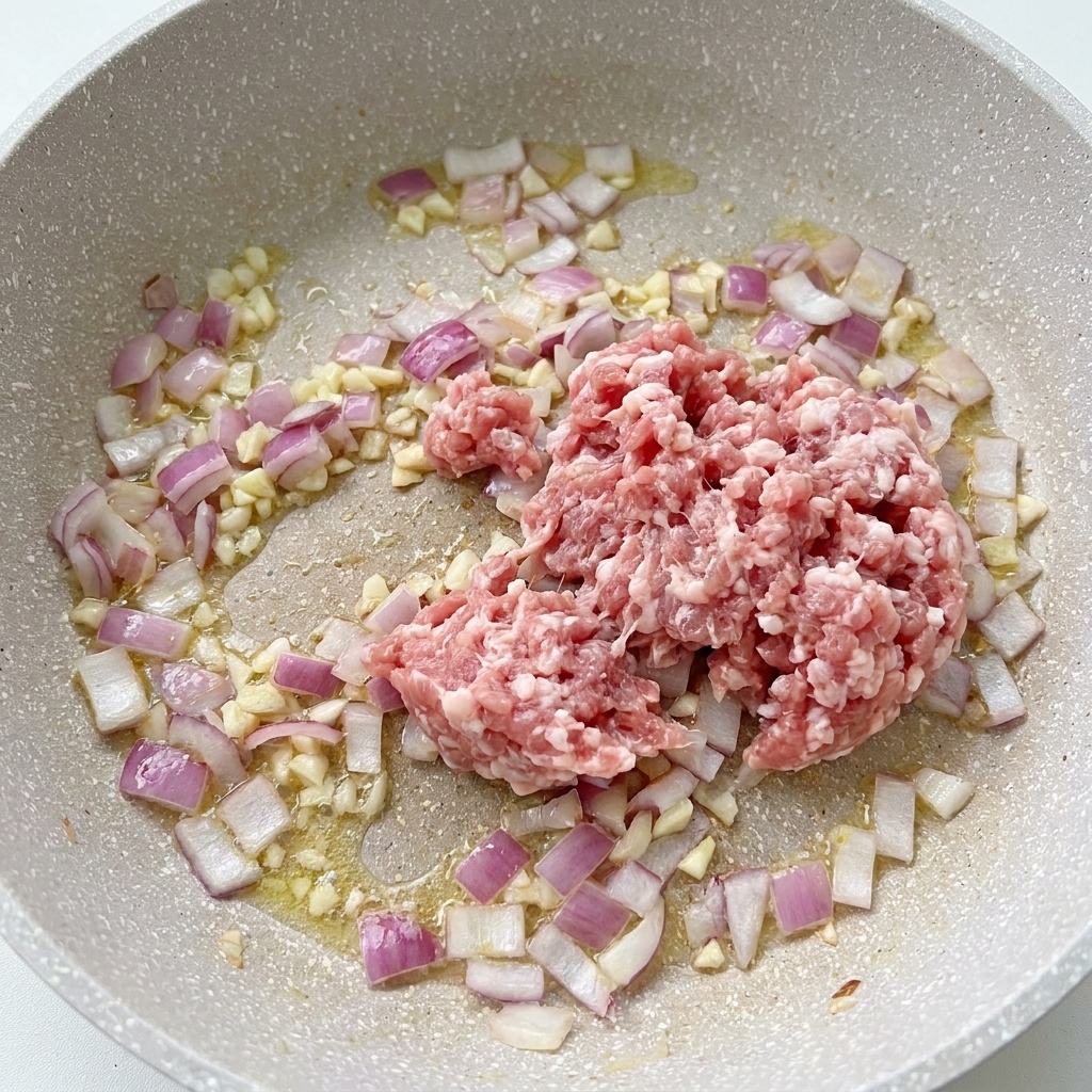 フライパンの中で、炒め中の玉ねぎやニンニクの横に生豚ひき肉が入っている様子。