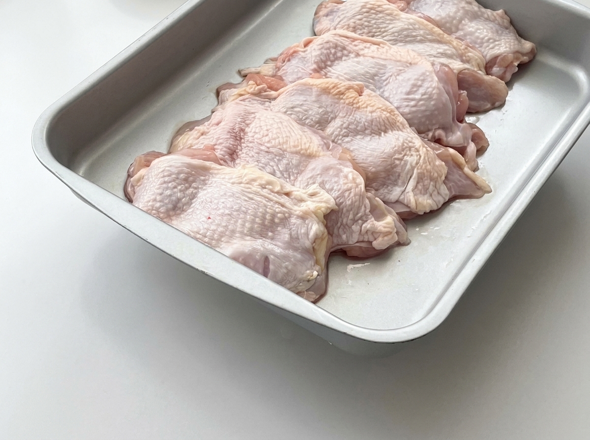 金属製のトレイに並べられた生の鶏もも肉と、その上に置かれた生姜のスライス。