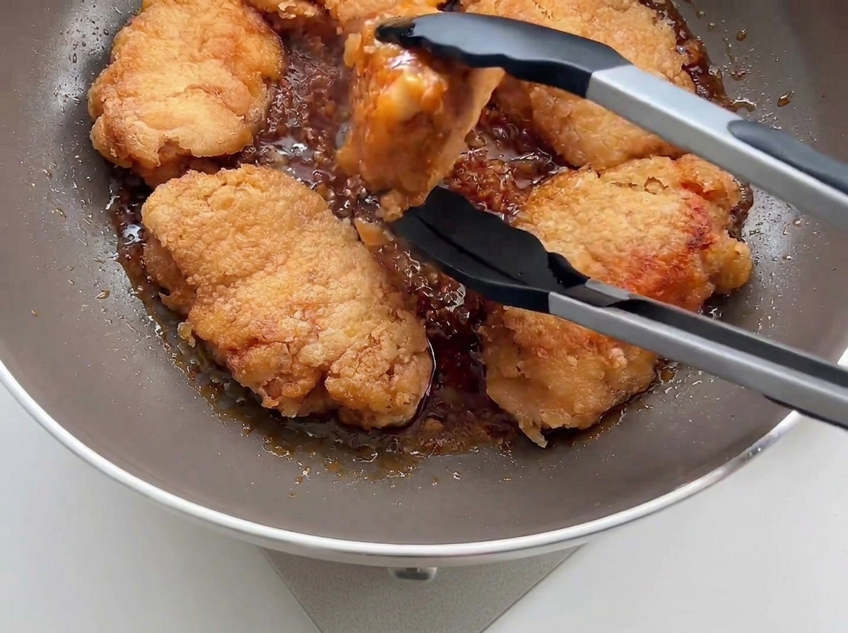 黒いトングを使って、カリカリのフライドチキンをガーリックソースの入ったフライパンに戻し入れている様子。