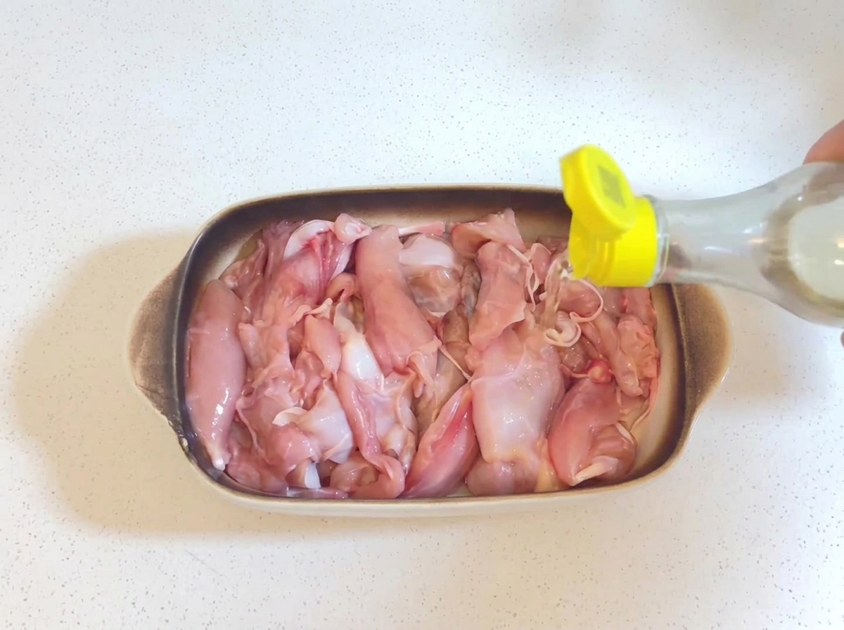 黄色いキャップの小さなボトルから、耐熱皿に入った生の鶏肉に透明な料理酒が注がれている様子。