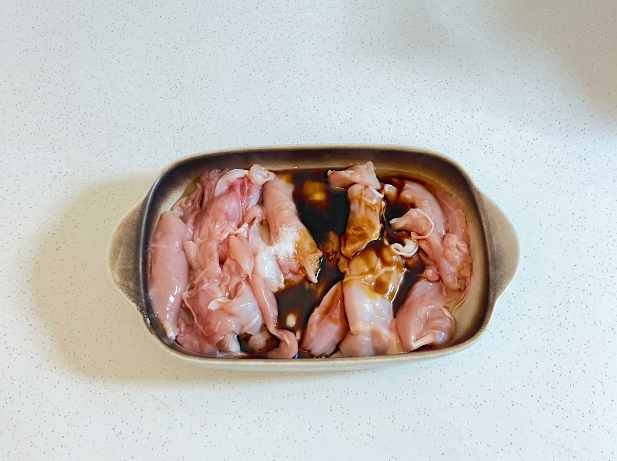 耐熱皿に入った生の鶏肉に、濃い色の調味液とサラダ油が注がれている様子。
