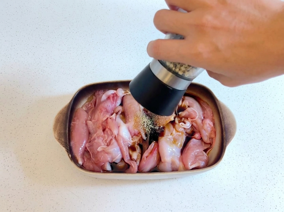 手に持ったペッパーミルで、皿の中でマリネされている生の鶏肉に挽きたてのコショウをふりかけている様子。