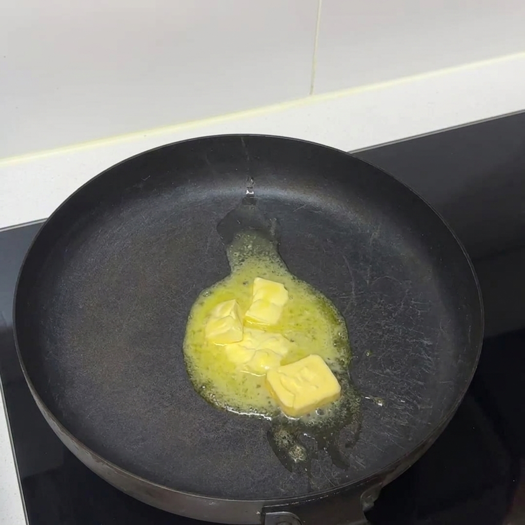 コンロに置かれた黒いフライパンの中で、バターの塊が溶けて液体になっている様子。