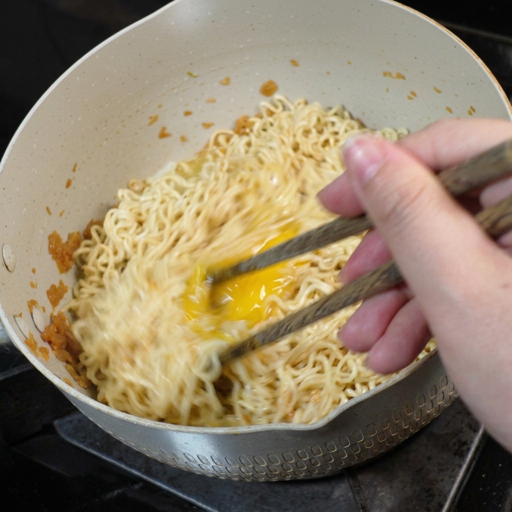木製の箸を使って、フライパンの中で卵が麺と一緒に混ぜられ、スクランブルされている様子。