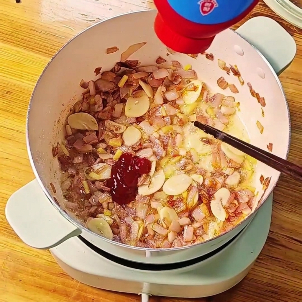 炒めた玉ねぎとニンニクが入った鍋に、赤いトマトペーストをチューブから絞り出している様子。