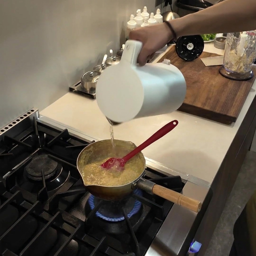 とろみがついて煮立っているフフの混合物が入った鍋に、白い容器から水を注いでいる様子。
