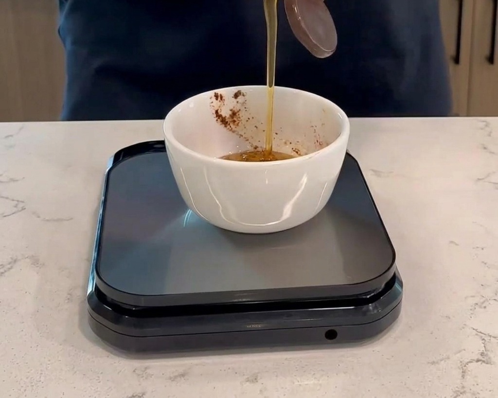 黒いデジタルキッチンスケールに置かれた小さな白いボウルに、ハチミツをプラスチックボトルから注ぎ入れ、濃い赤色のソースと合わせている様子。