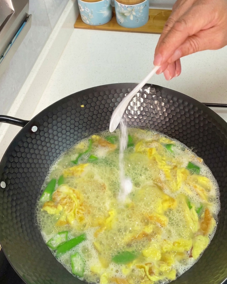 煮込んでいるオクラと卵のスープに、スプーンで塩を加える様子。