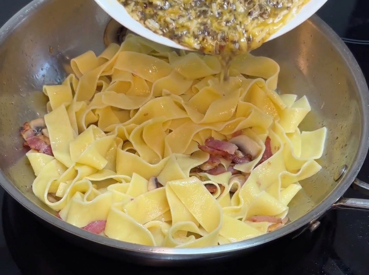 白いお皿から、平打ちパスタの入ったステンレス製のフライパンに、あらかじめ作っておいた黒トリュフカスタードソースが注がれている様子。