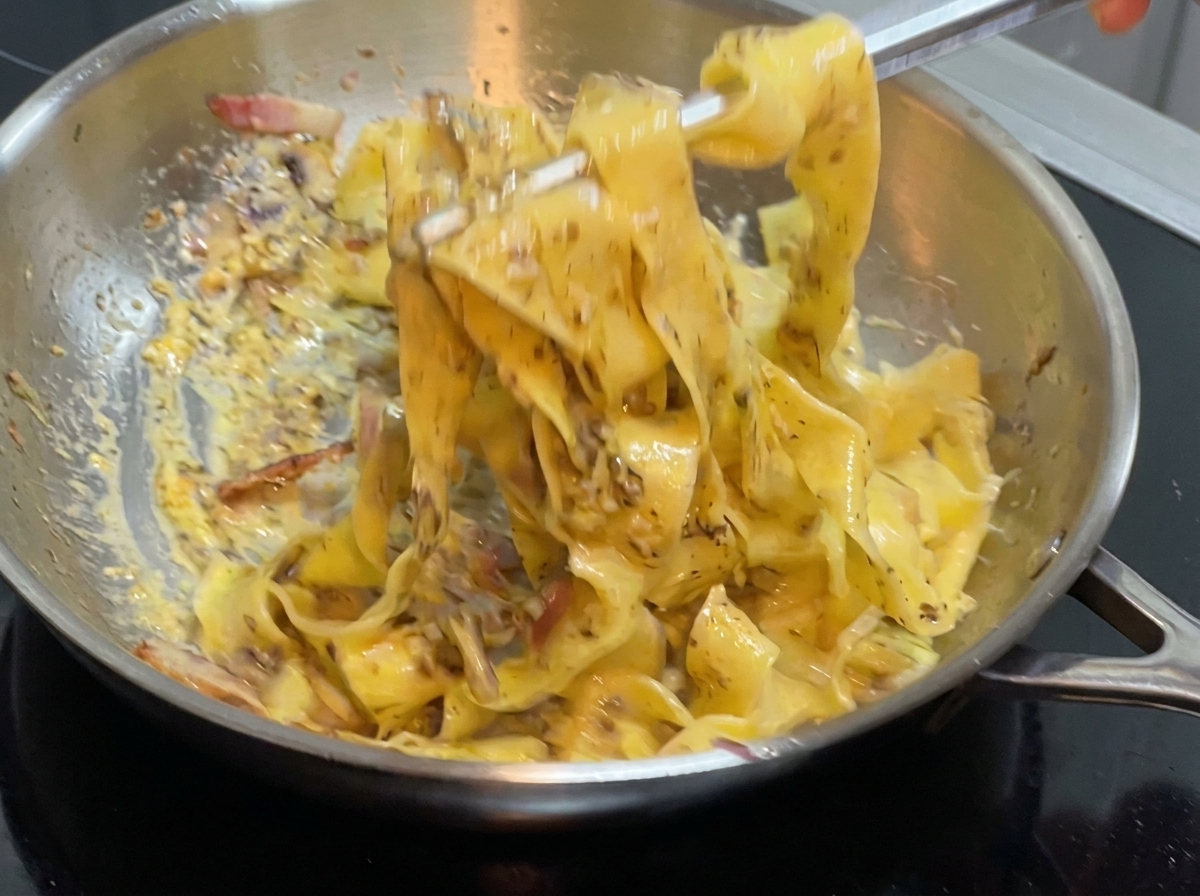 コンロの上のステンレス製フライパンの中で、トングを使って平打ちパスタとソースを混ぜ合わせている様子。