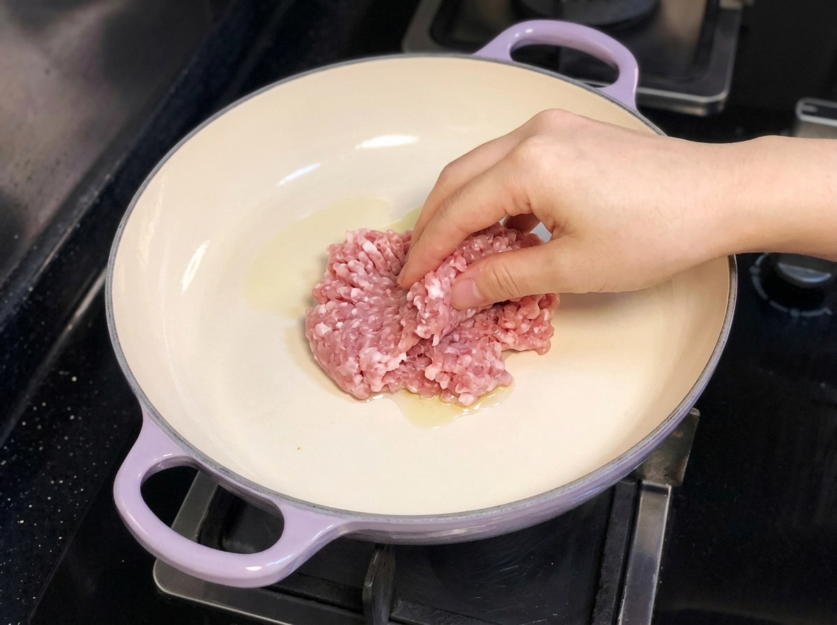 紫色の取っ手のついたフライパンにオリーブオイルを熱し、手で豚ひき肉を入れている様子。