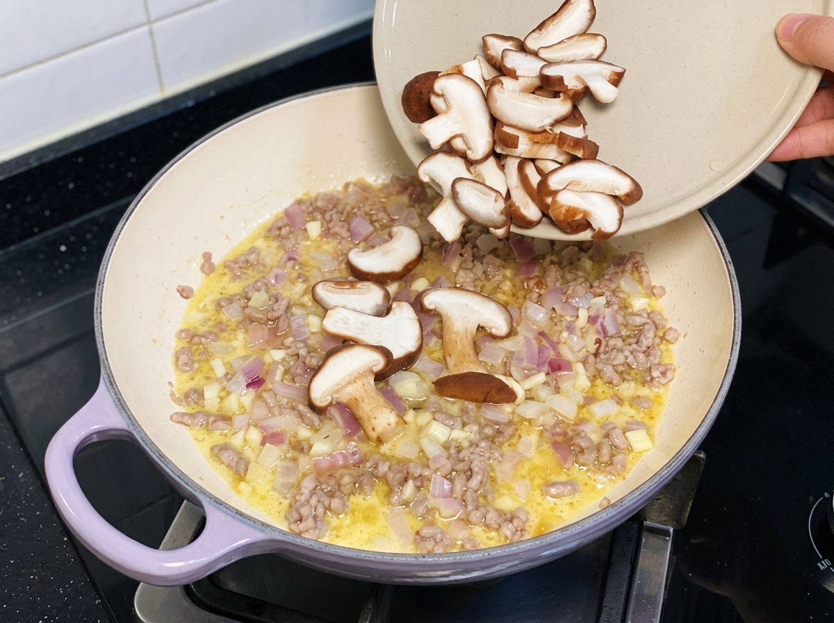 お皿から、豚ひき肉と玉ねぎを炒めているフライパンへスライスした生椎茸を入れている様子。