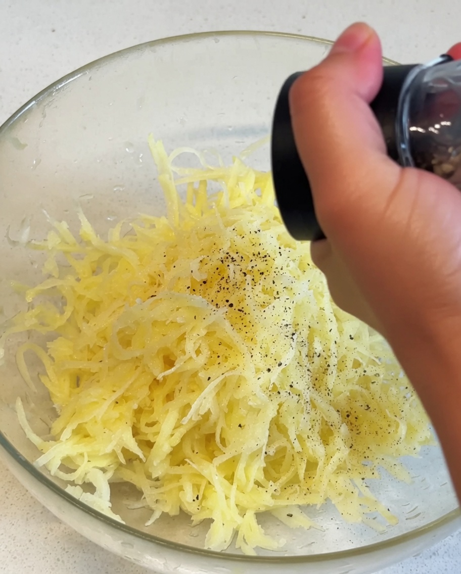 ガラスボウルに入った千切りじゃがいもに、手動ミルで黒こしょうが振りかけられている様子。