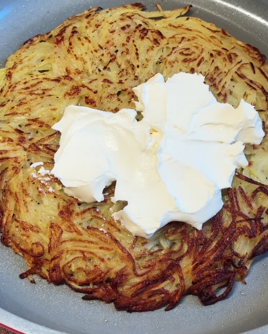 熱々のカリカリなじゃがいものパンケーキ全体に、白いクリームチーズが乗せられ、滑らかに伸ばされている様子。