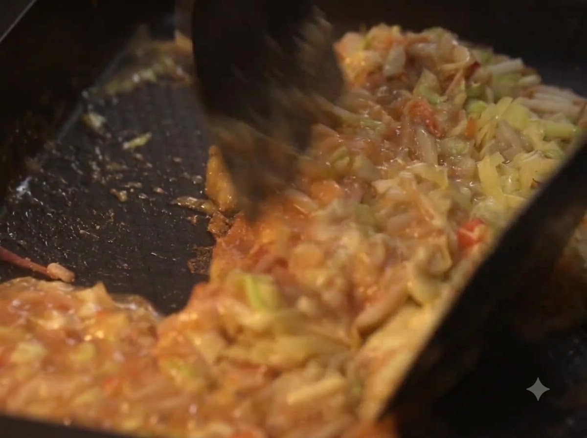 熱いプレートの上で、ヘラを使って調理中の野菜と液体生地を混ぜ合わせている様子。