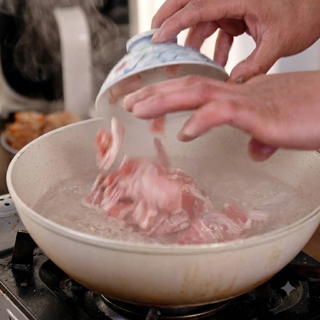 小さなセラミックボウルから切った生肉の豚バラ肉を、沸騰している鍋へ手で落とし込んでいる様子。