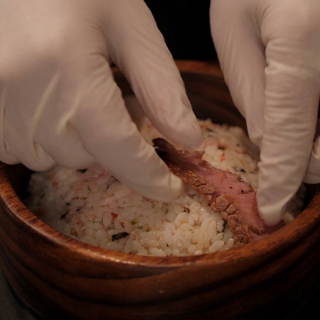 手袋をした手が、調理したステーキのスライスを味付けされたご飯の丼の上に並べている様子。