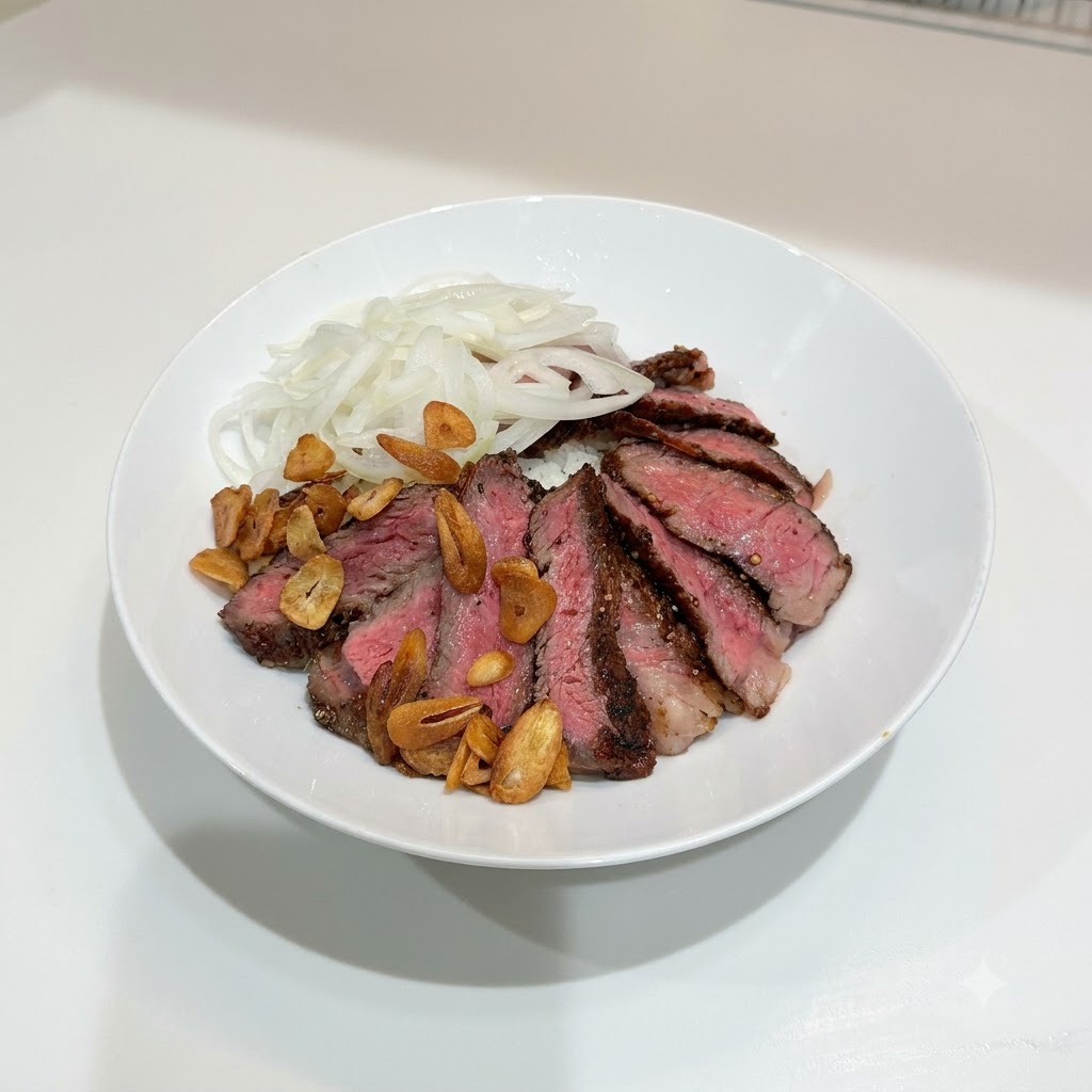 白い丼にご飯、スライスしたステーキ、玉ねぎが盛られ、その上にきつね色のガーリックチップが散りばめられている様子
