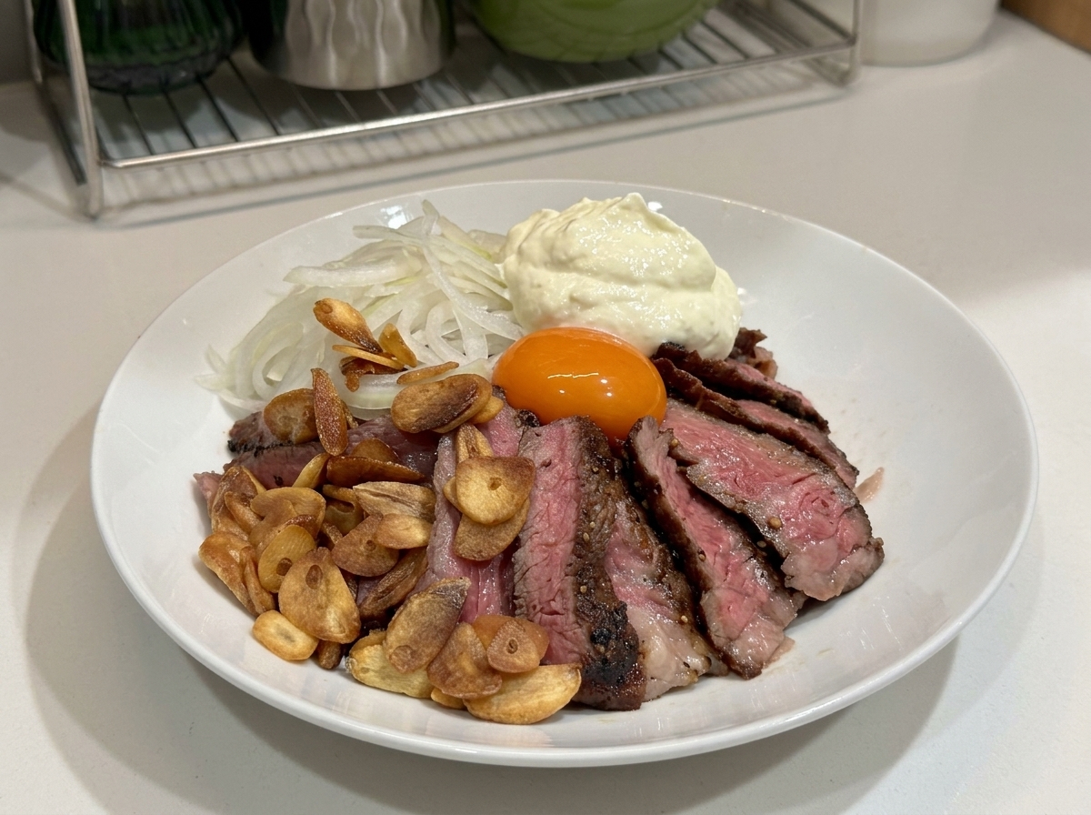 スライスしたステーキ、ガーリックチップ、玉ねぎ、卵黄、そして大盛りのホイップクリームがトッピングされた白い丼