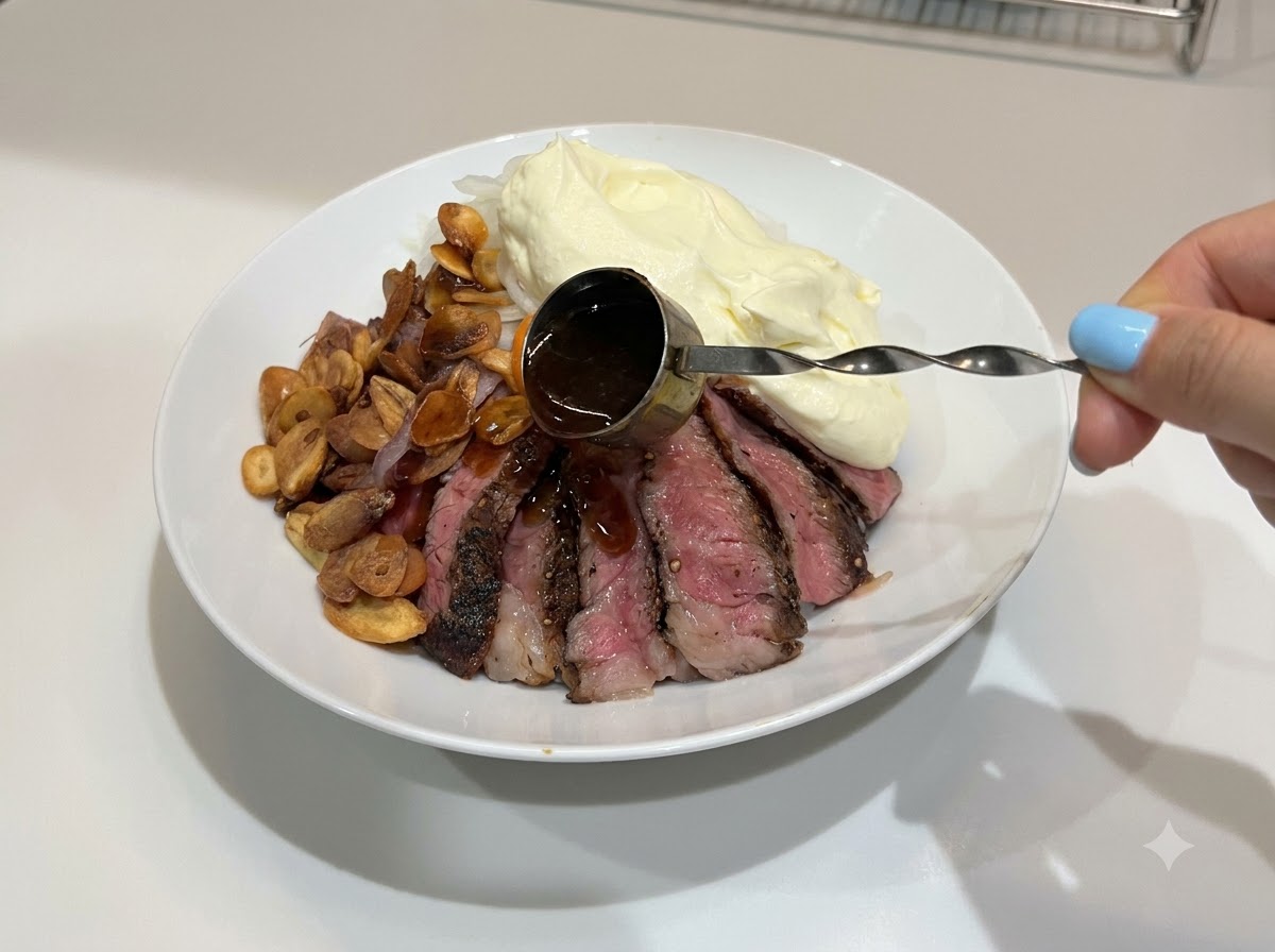 小さな計量カップを持った手で、完成したステーキライスの丼の上に濃い色のテリヤキソースを回しかけている様子