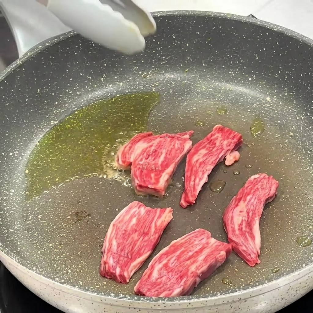 フライパンで調理中のステーキ肉に、塩と黒こしょうを振っている様子。