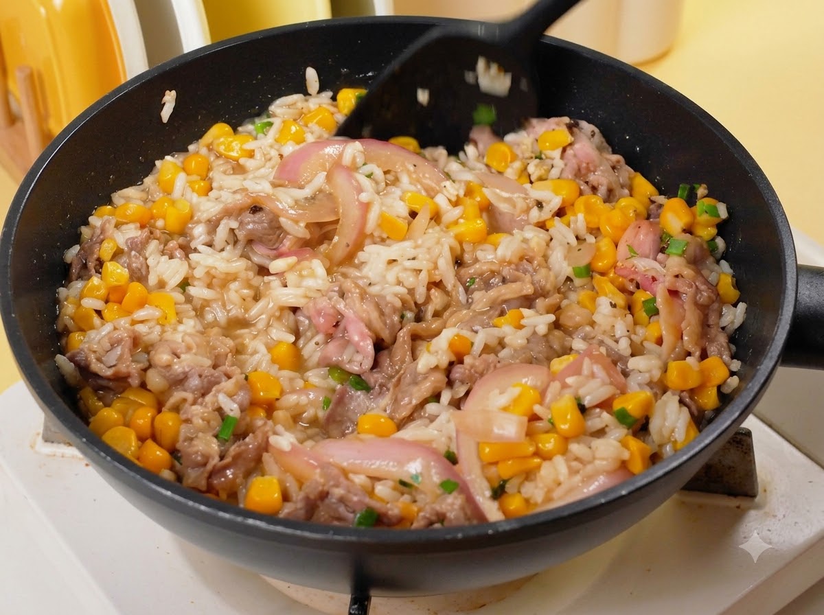 黒いスパチュラを使って、フライパンの中でご飯、牛肉、コーン、玉ねぎを混ぜ合わせながら炒めている様子。