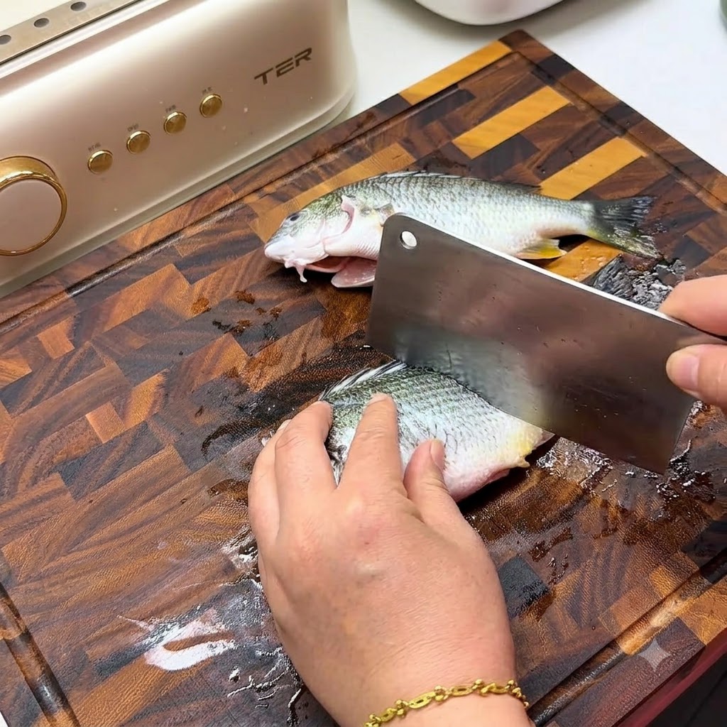 木製のまな板の上で、大きな中華包丁を使って新鮮な魚の側面に斜めの切れ目を入れている様子。