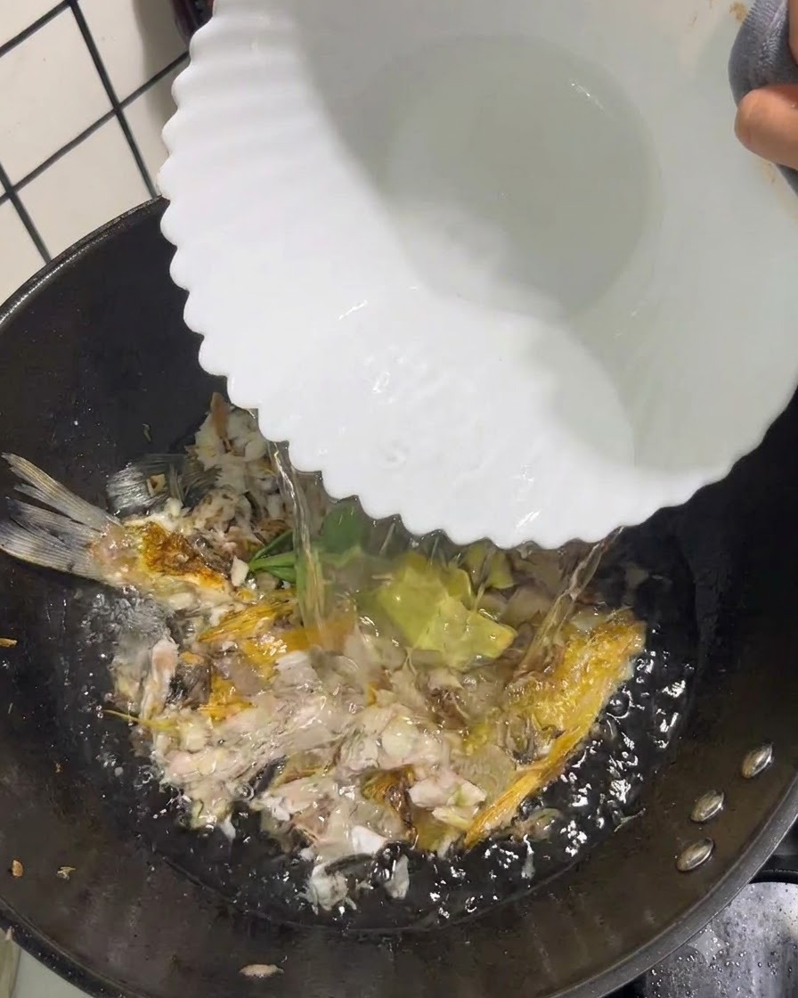 砕いた魚と香味野菜の入った中華鍋に、白いボウルから透明な熱湯を注ぎ入れている様子。