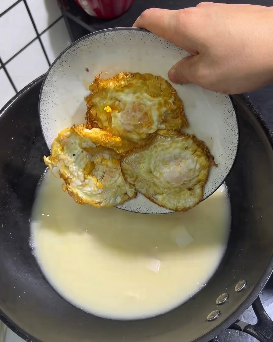 白い皿から黄金色の目玉焼き3個を、煮立っているスープの鍋に入れている様子。
