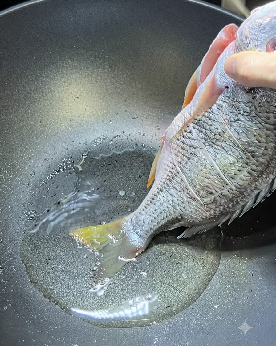 片手で丸ごとの生黄鯛の上半身を持ち、その尾がフライパンの熱い油に触れている様子。