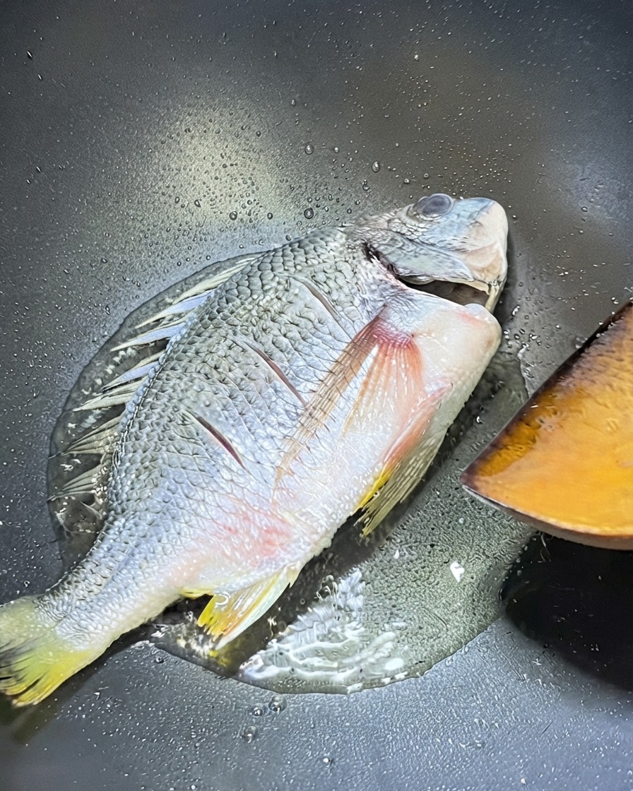 フライパンで焼かれている黄鯛と、裏返すために近くに置かれた木製スパチュラ。