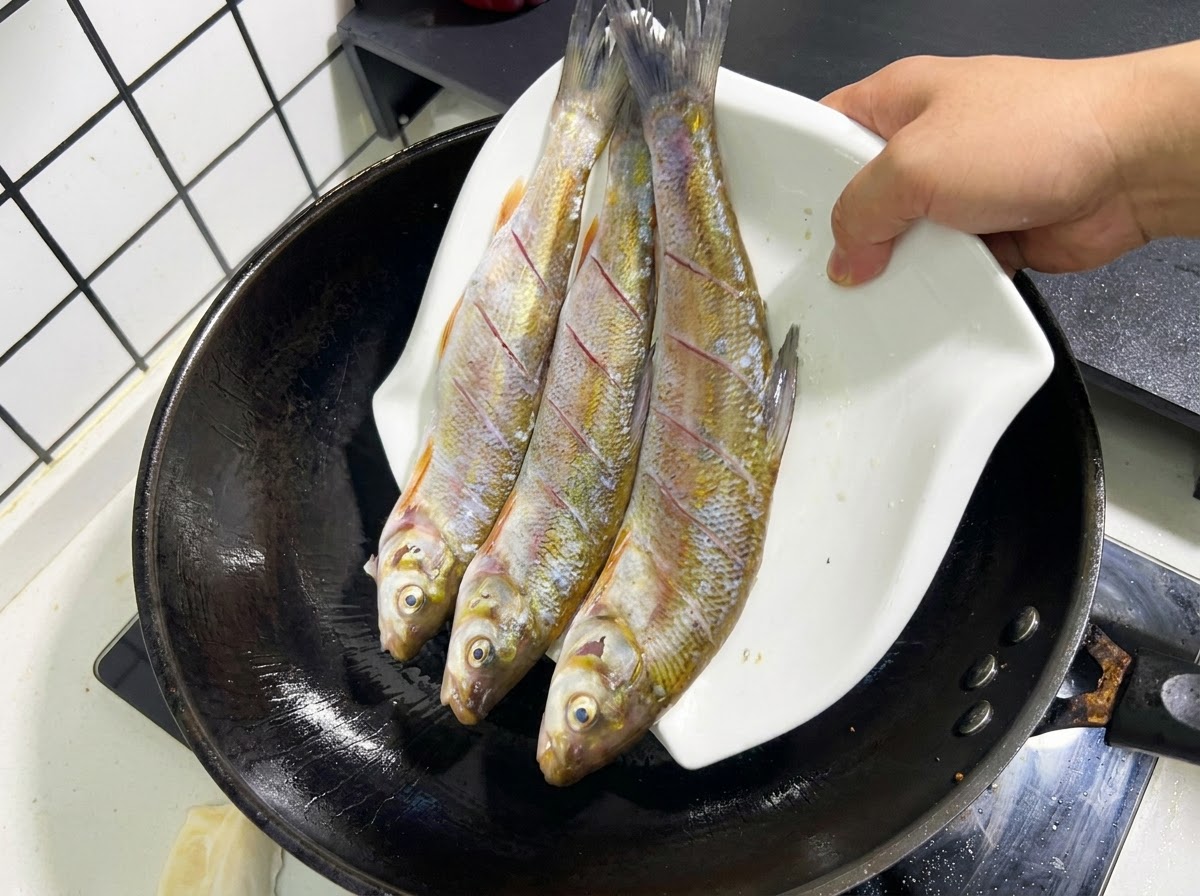 白い皿にのった3尾の下処理済みの魚を、熱した中華鍋の油へ慎重に滑り込ませている様子。